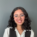 Smiling woman with glasses and curly hair stands against a gray background, wearing a white blouse and dark vest, conveying a professional and approachable vibe.