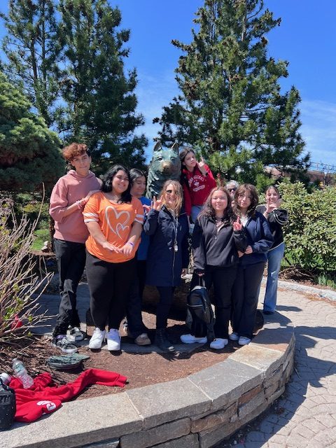 A group of approximately eight 8th graders stands together outdoors on a paved walkway next to Jonathan the Husky statue. The group is gathered closely, with several people in the front and others slightly behind.