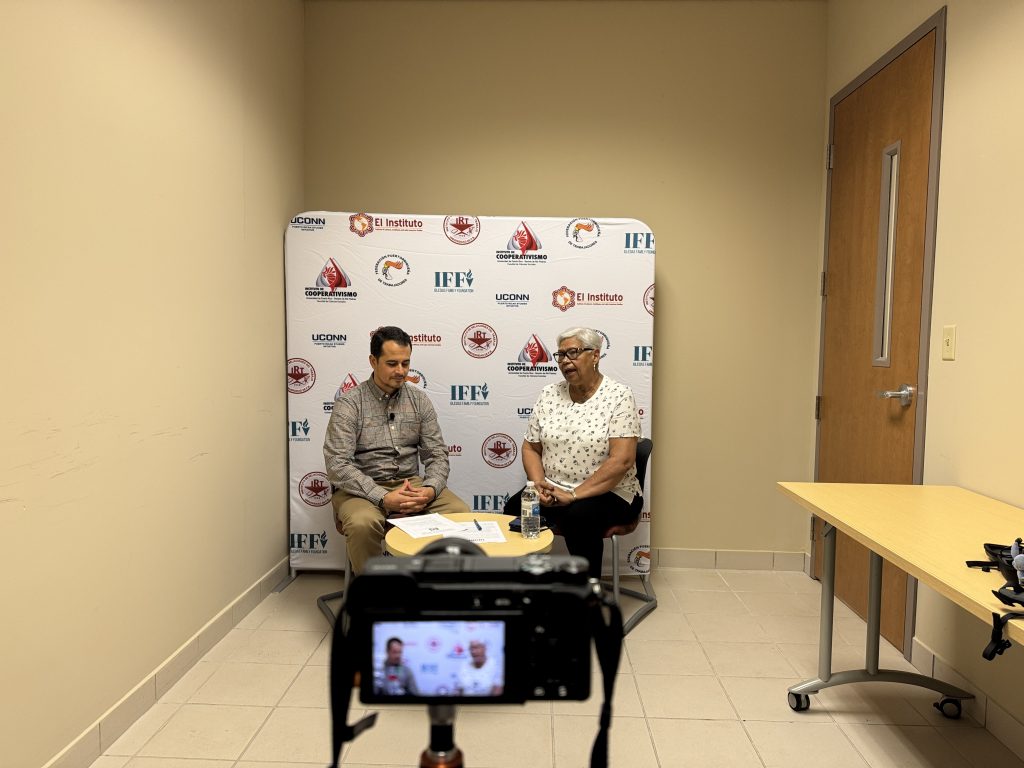 A camera on a tripod is positioned in the foreground, recording two seated adults in a small room. The two people sit on chairs facing the camera with a small round table between them holding papers, a pen, and a water bottle. Behind them is a step-and-repeat backdrop displaying multiple logos, including UConn, El Instituto, IFF, and Cooperativismo. 