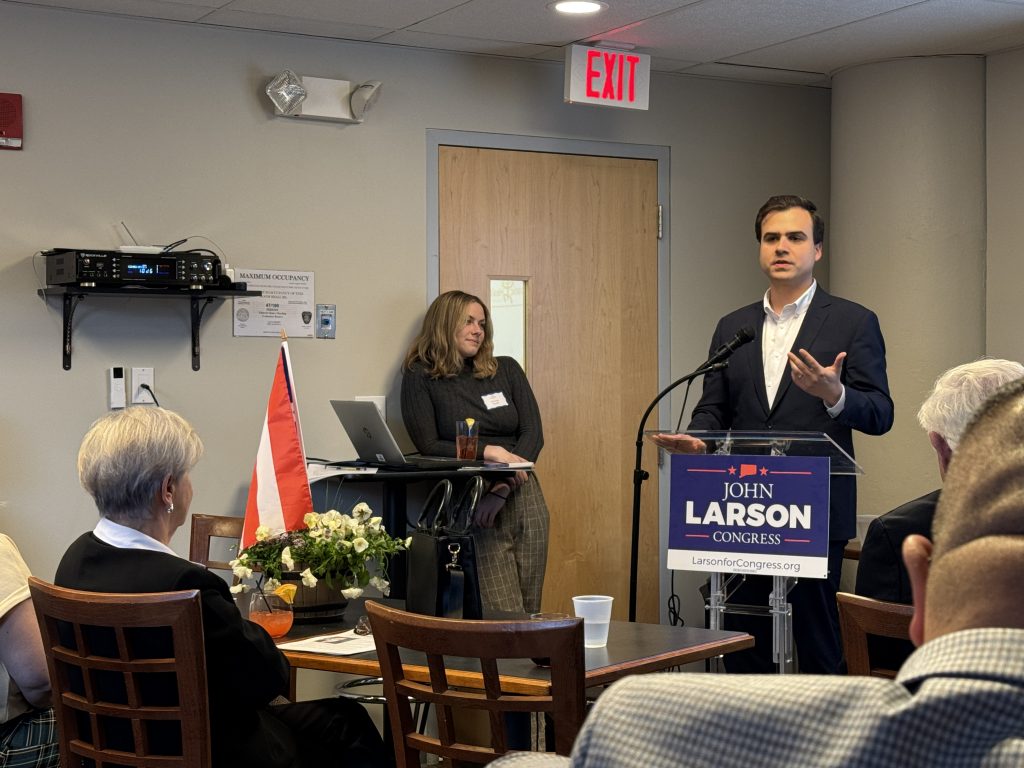 Representative John Larson stands at a clear podium speaking into a microphone in a small indoor event space, with an audience seated at tables in the foreground. 