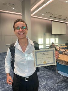 Graduate student Beatriz Torres wearing a white button-down shirt and black pants stands indoors holding a certificate labeled “Certificate of Participation.”