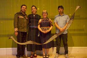 Four adults stand side by side in a gallery, viewed through a curtain of vertical hanging threads with small colorful attachments. A long, black-and-white patterned woven textile curves across the foreground at waist height.