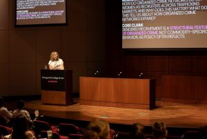 A woman stands at a podium, presenting to an audience at a conference, with a projector screen behind her.
