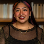 Graduate Student Vania with red highlights wearing a black top, smiling and looking directly at the camera.